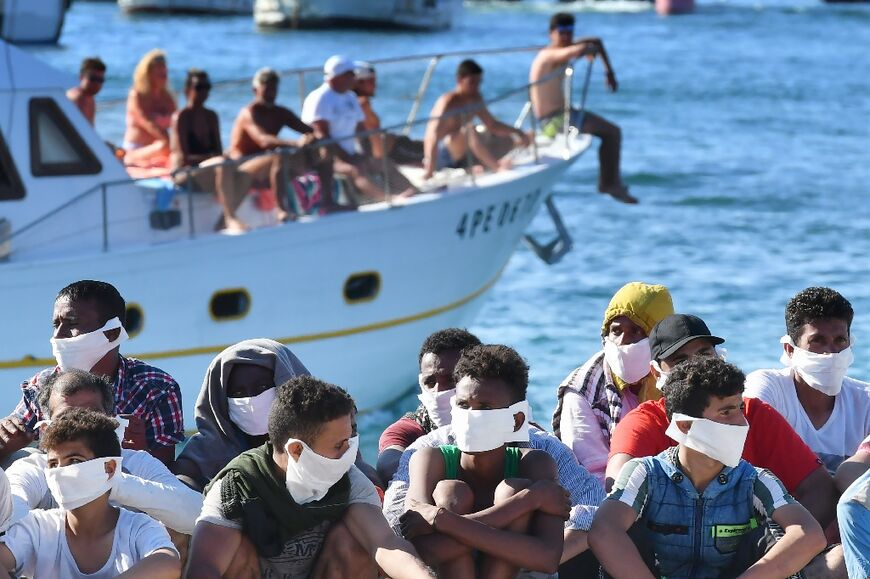 Migrants rescued by Italy's coast guard sit as a boat with tourists enters a harbor on the island of Lampedusa