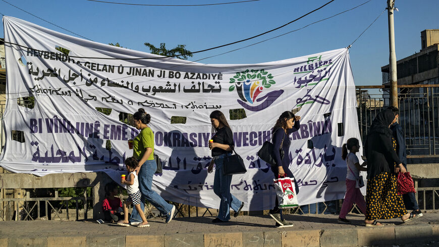 Syrian Kurdish women walk past an election campaign banner in the northeastern Syrian city of Qamishli on June 6, 2024. 