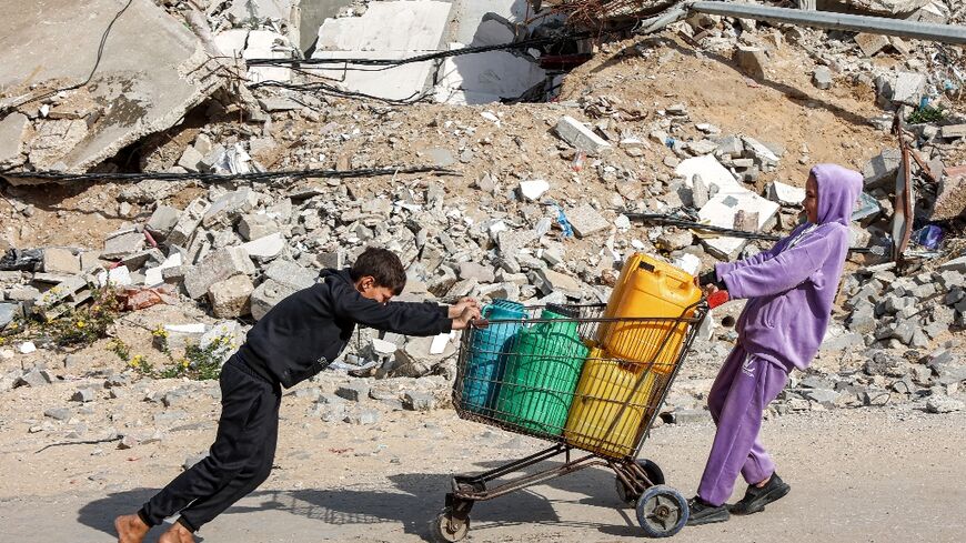 A girl pulls while a boy pushes a shopping-cart loaded with filled-up water containers past a mound of rubble and debris in Gaza City