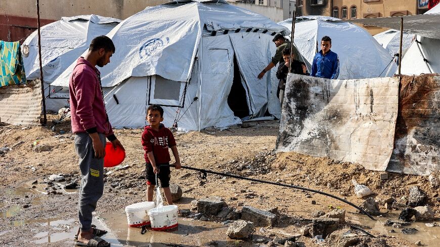 A youth speaks to a boy filling up buckets with water from a hose near tents at a shelter for displaced Palestinians in Gaza City