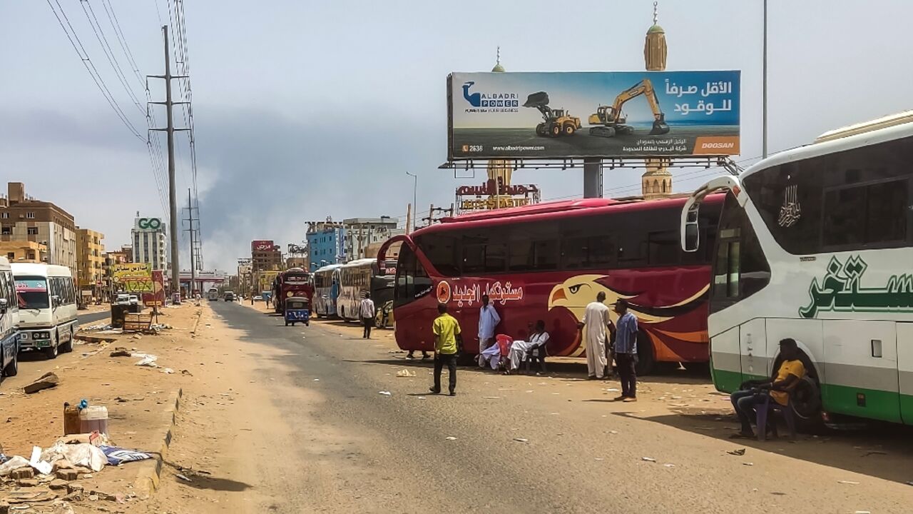 Smoke billows in the distance as people wait next to passenger buses in Sudan's capital Khartoum