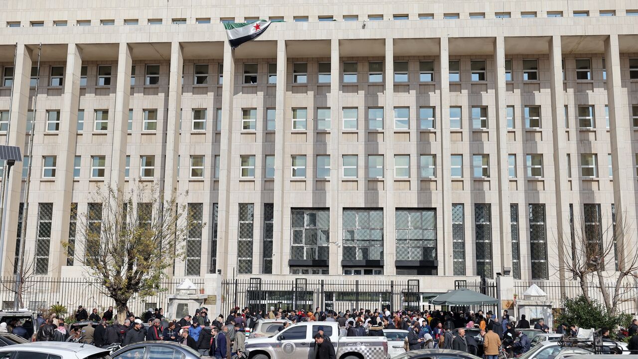 Vehicles move past as people queue outside the main headquarters of the Central Bank of Syria to exchange US dollars at prices lower than the black market.