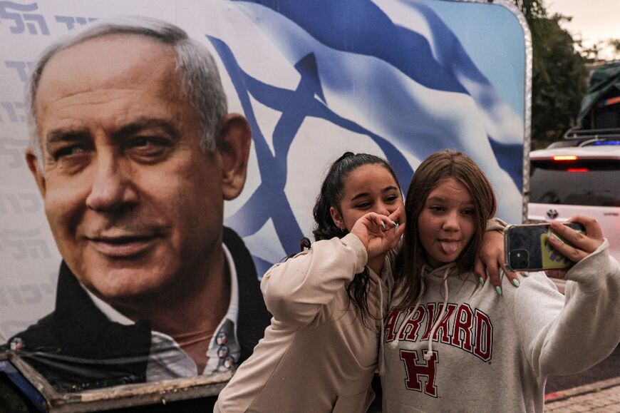 Two girls pose for a selfie before a camper wagon showing a Likud party electoral banner depicting its leader Benjamin Netanyahu, on October 23, 2022
