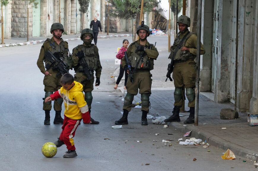 A Palestinian boy plays football on the street next to Israeli security forces on patrol in the city center of Hebron in the occupied West Bank, on November 18, 2022