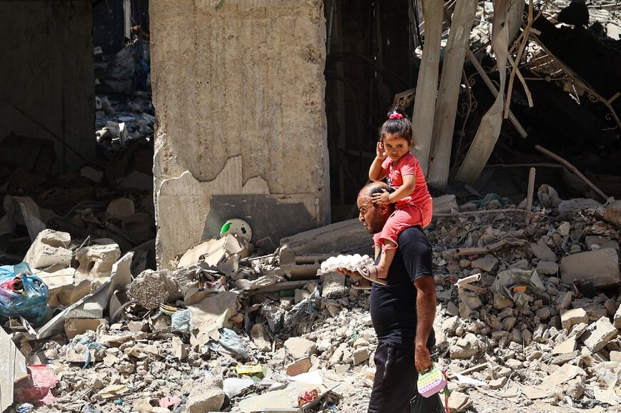 A Palestinian carrying a child on his shoulders clutches a carton of eggs as he walks past a destroyed building in the Gaza city of Khan Yunis