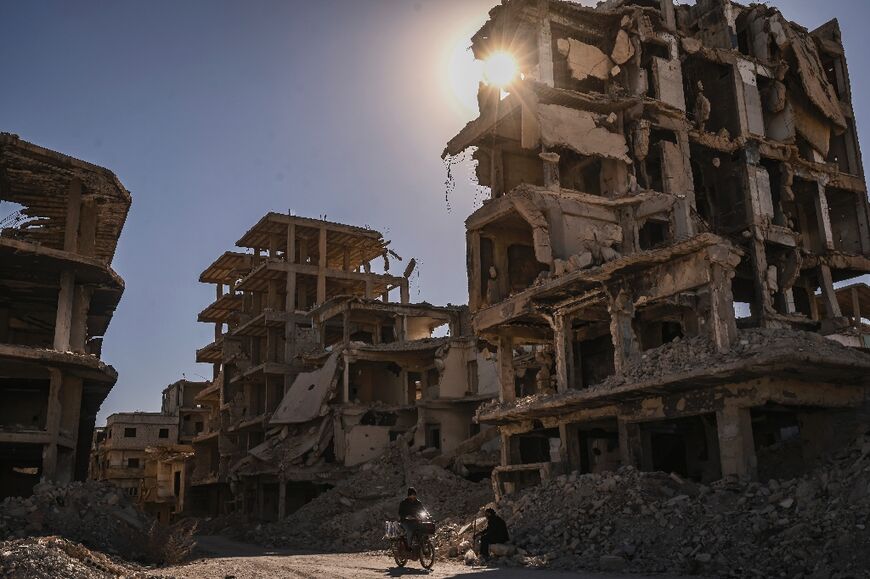 A man rides a motorcycle past destroyed buildings as another looks on at the Yarmuk camp for Palestinian refugees in the south of Damascus